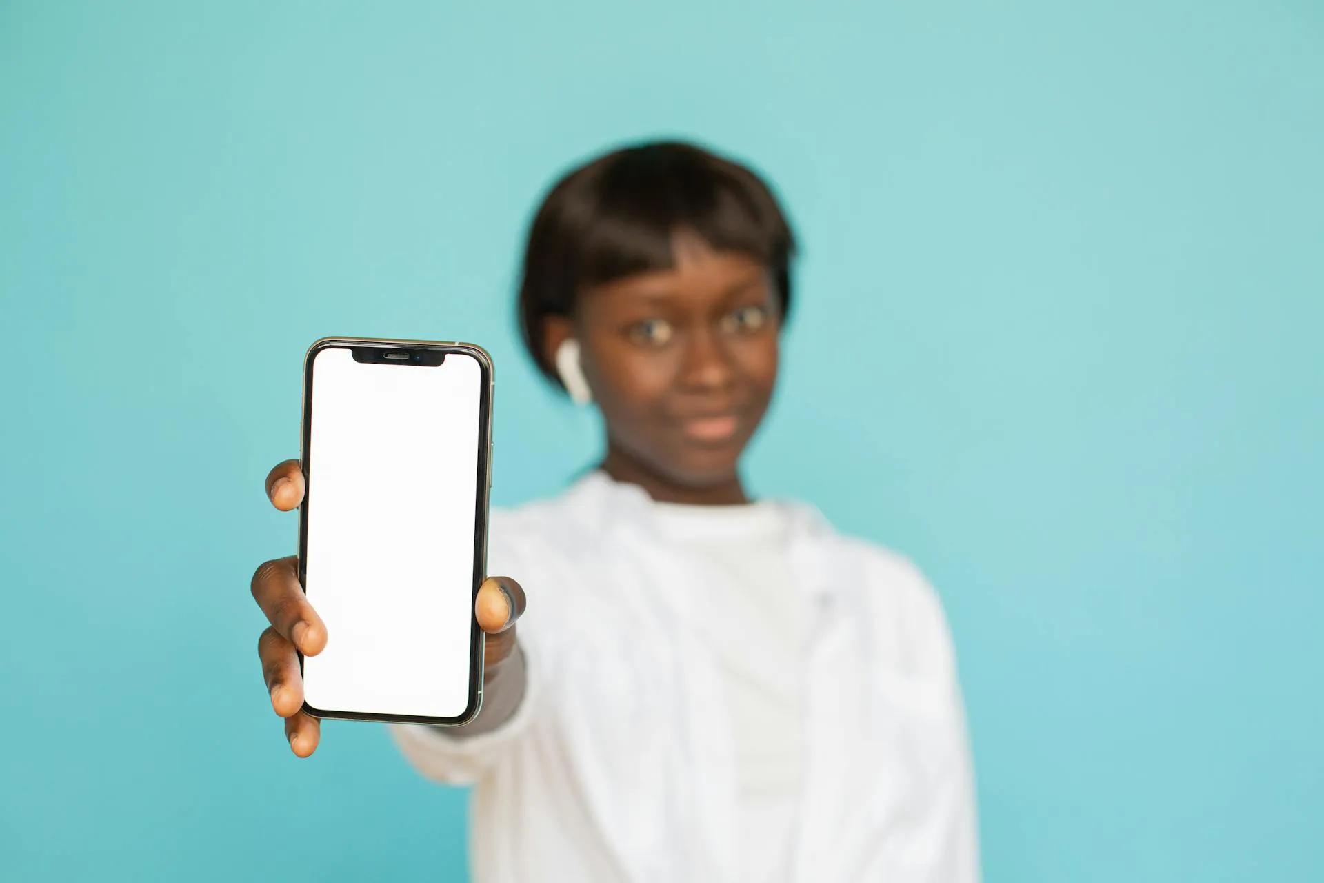 Une personne aux cheveux courts, portant une chemise blanche et des écouteurs sans fil, tient un smartphone avec un écran blanc vierge en direction de la caméra sur un fond bleu clair - idéal pour ceux qui recherchent des emplois d'étudiants dans le domaine du tutorat en ligne ou des moyens de gagner de l'argent en ligne en tant qu'étudiant.