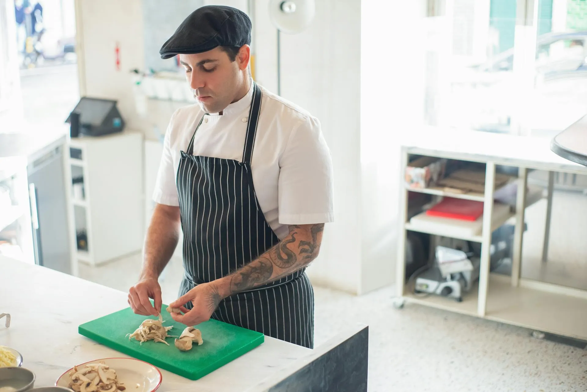 Un chef cuisinier portant une casquette noire, une chemise blanche et un tablier rayé coupe des champignons sur une planche à découper verte dans une cuisine moderne et lumineuse - une inspiration parfaite pour les idées de revenus en ligne des étudiants.