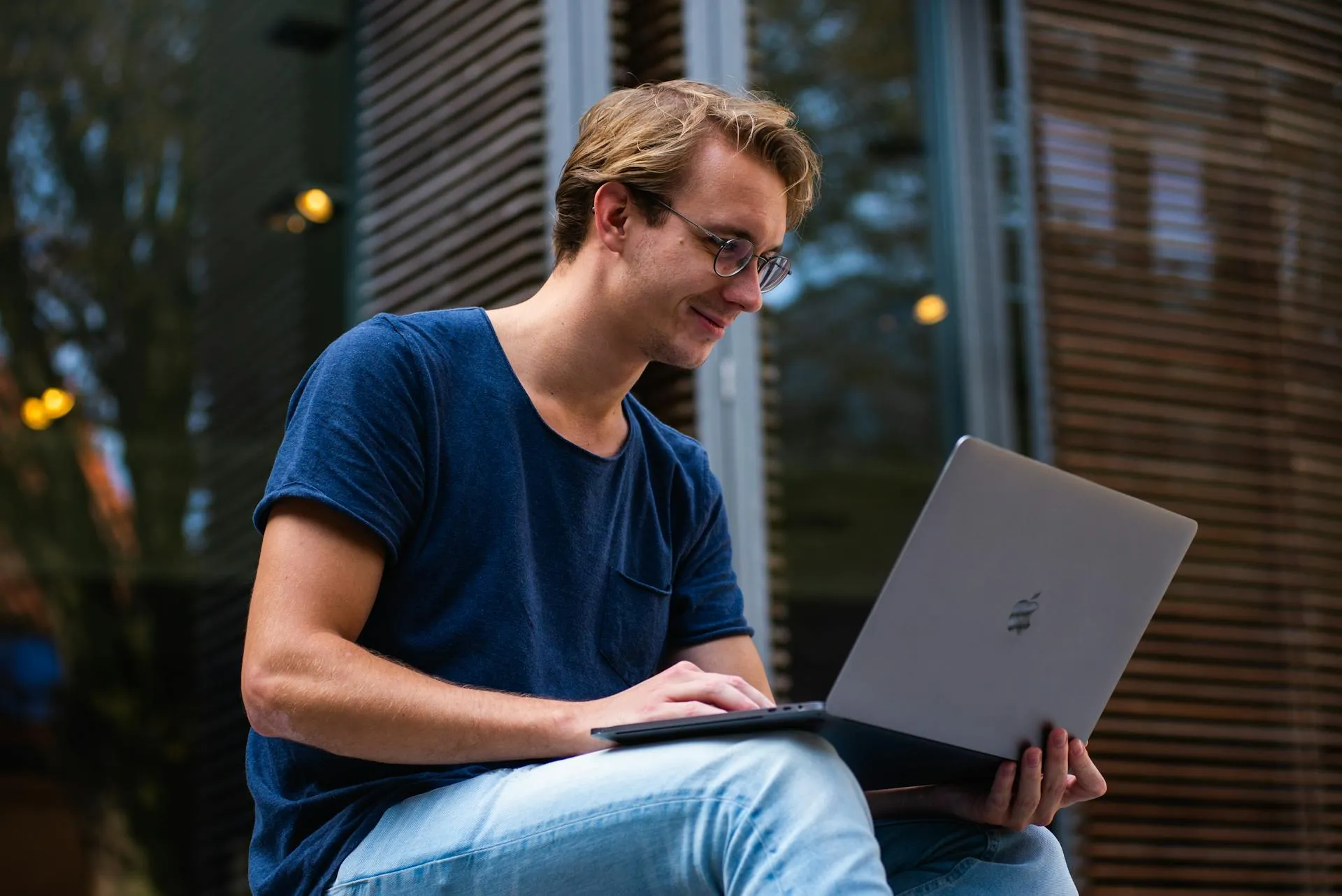 Un jeune homme portant des lunettes et un t-shirt bleu est assis à l'extérieur, souriant tout en travaillant sur un ordinateur portable - peut-être en explorant les plateformes de freelancing 2025 pour gagner de l'argent en ligne en tant qu'étudiant. De grandes fenêtres avec des stores en bois reflètent les arbres à l'arrière-plan.