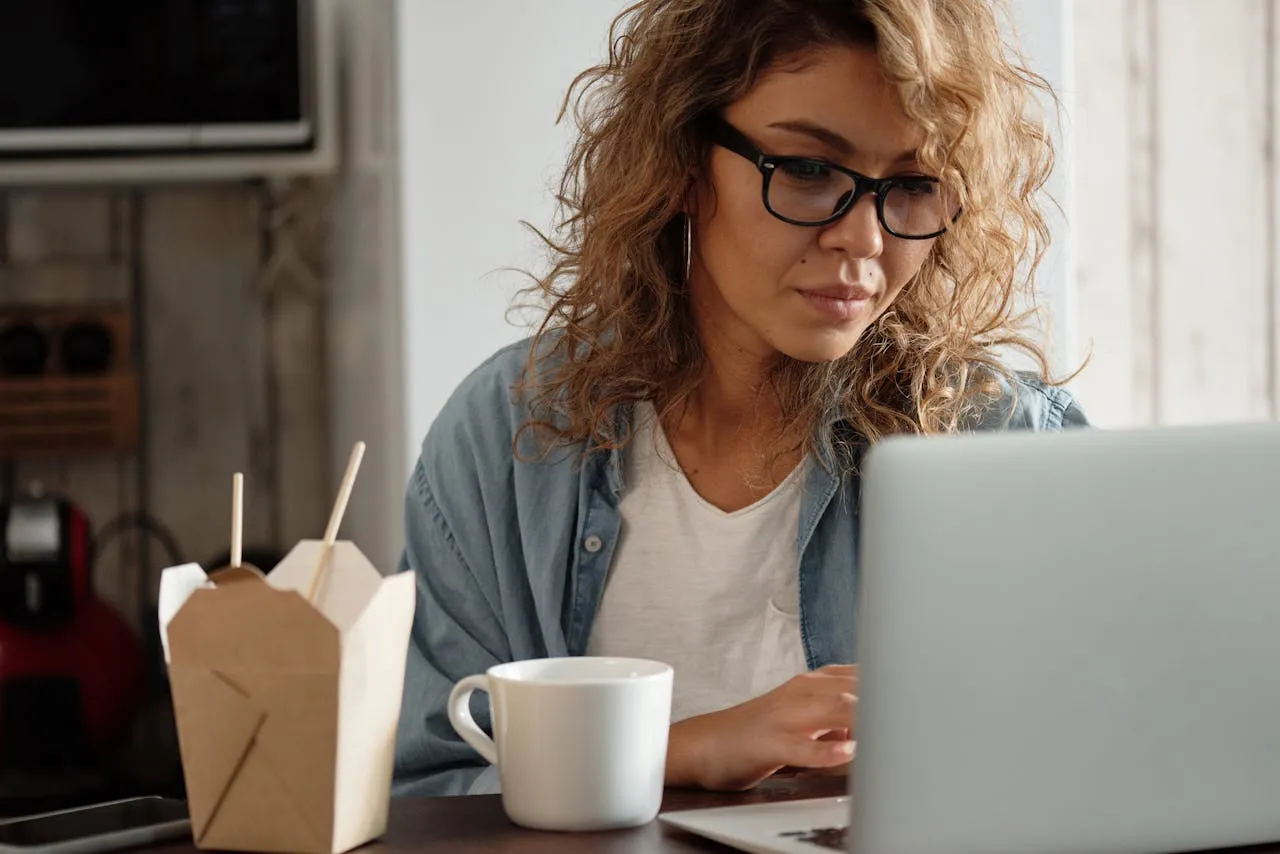Une femme aux cheveux frisés et aux lunettes est assise à une table, devant un ordinateur portable. Devant elle se trouvent un récipient à emporter avec des baguettes et une tasse à café. Elle semble concentrée, explorant des idées de revenus en ligne pour les étudiants dans un environnement intérieur.