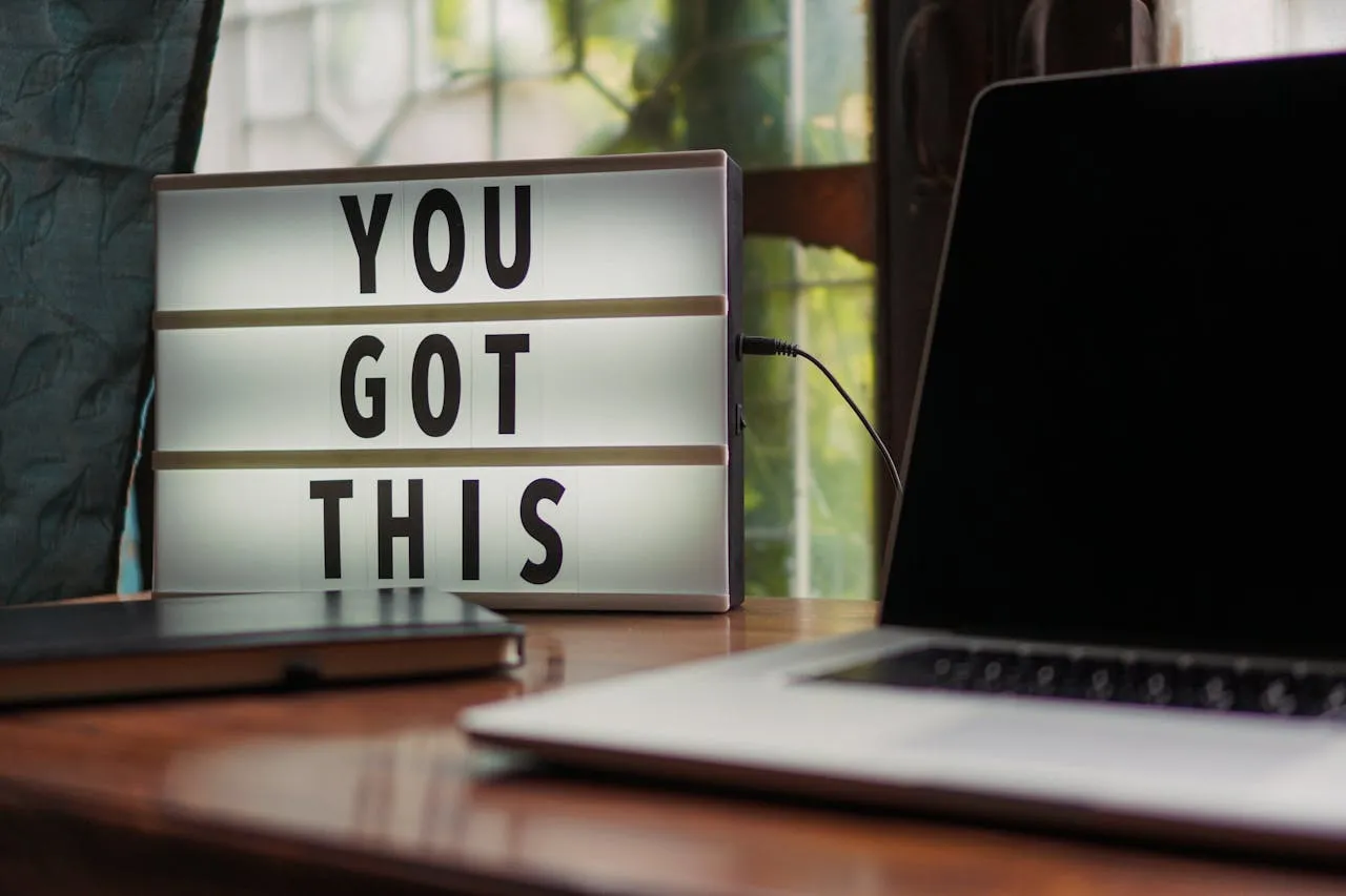Un caisson lumineux posé sur un bureau en bois affiche "YOU GOT THIS" à côté d'un cahier fermé et d'un ordinateur portable ouvert, mettant en évidence une configuration lumineuse et inspirante pour ceux qui recherchent des emplois de test de produits à distance ou qui cherchent à gagner de l'argent en ligne en tant qu'étudiant.